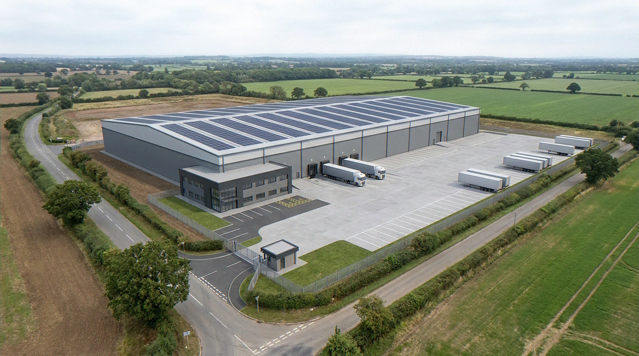 Aerial photomontage CGI of the same UK site after construction, with a build-to-suit logistics warehouse, rooftop photovoltaic array, full row of dock-levellers with articulated HGVs reversed in, fifty-metre concrete yard, trailer parking, gatehouse with ANPR lane, two-storey anthracite office frontage, and palisade perimeter