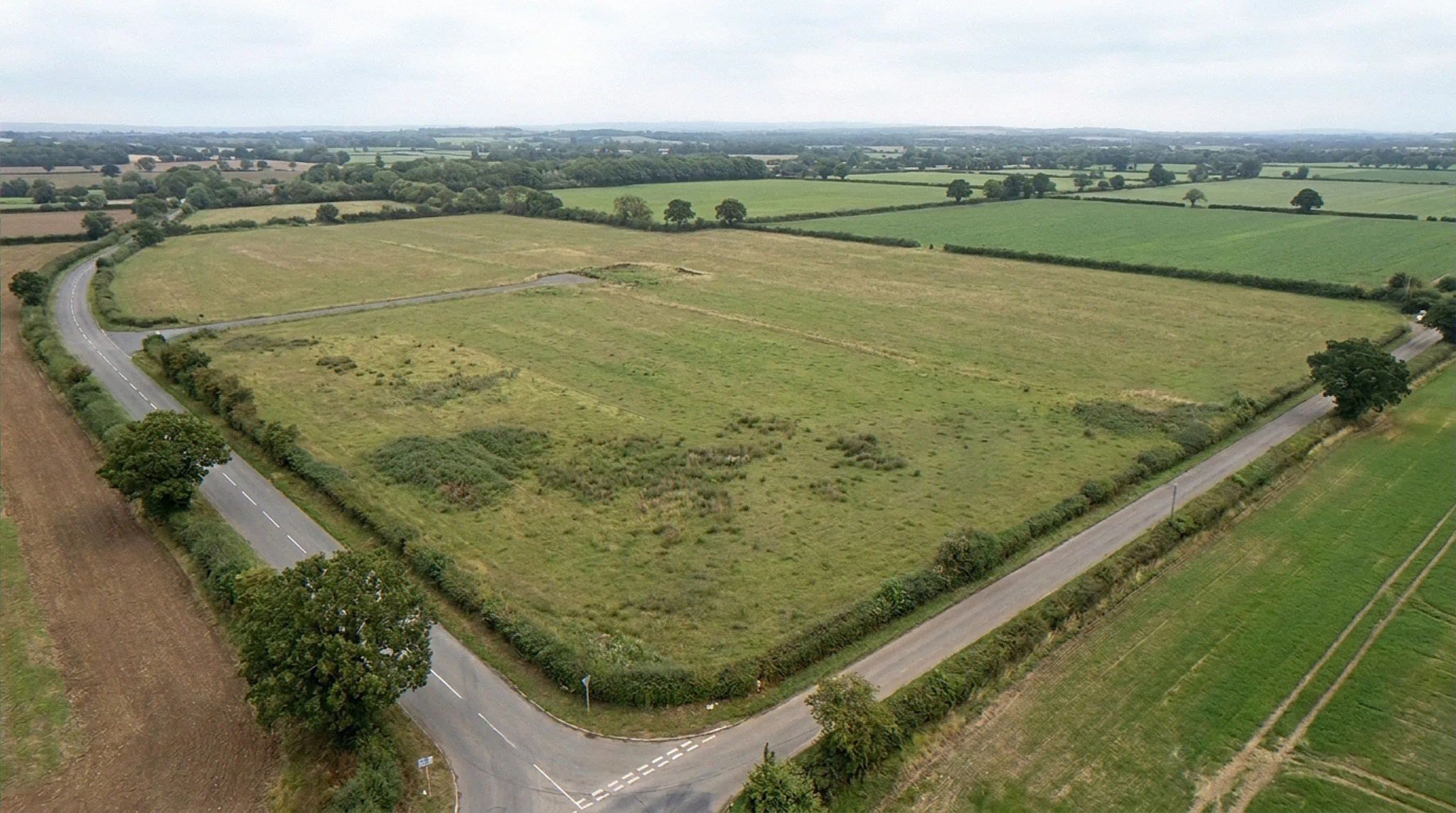 Aerial CGI of a flat UK greenfield agricultural site before construction, with country B-road, perimeter hedgerow, mature oak trees, and neighbouring fields under overcast British daylight