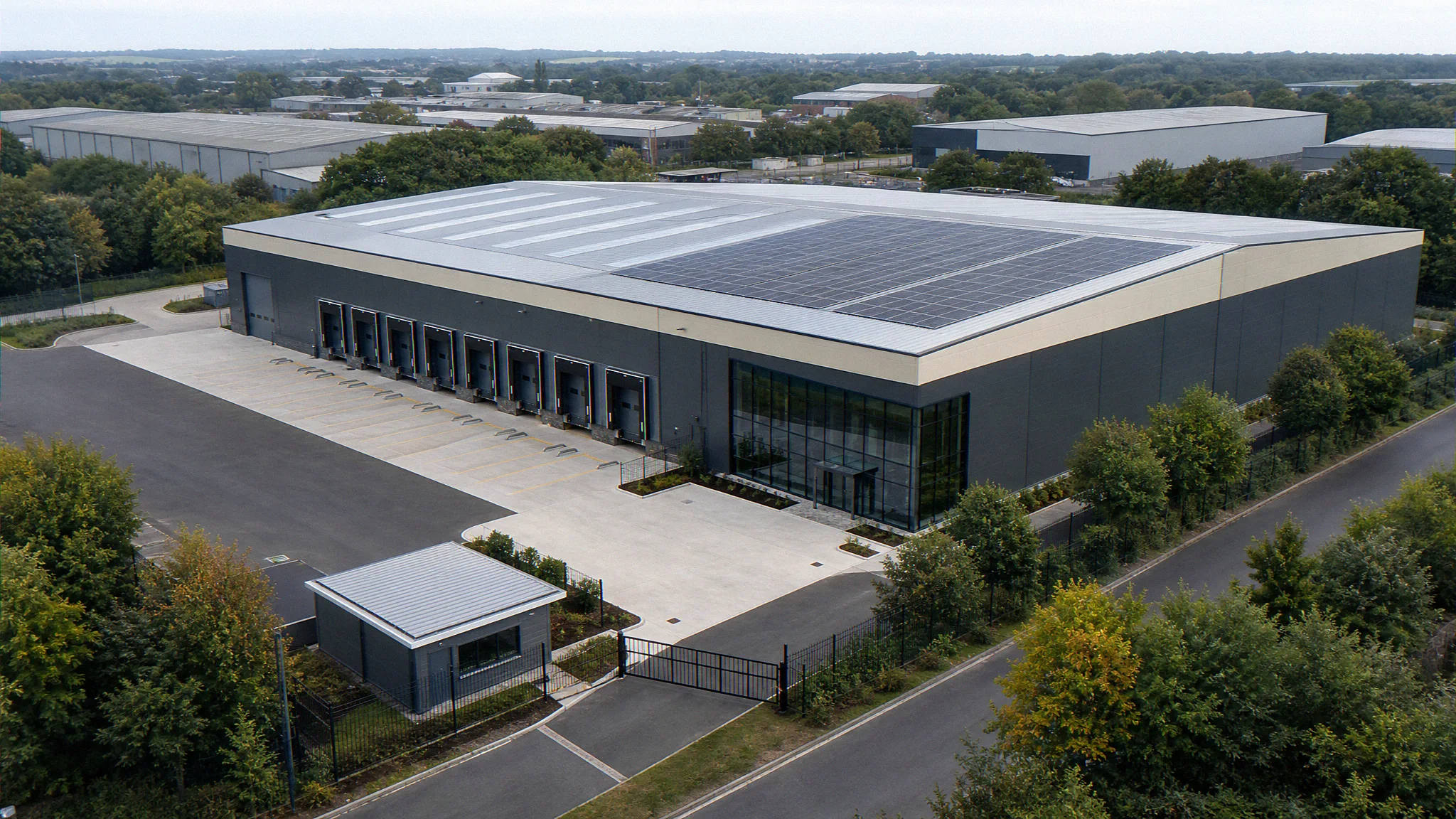 Wide aerial three-quarter architectural CGI of a UK industrial warehouse refurbishment scheme showing the recladded envelope, new rooflights, roof-mounted solar PV array, refreshed yard, and dock face from a coordinated 3D model