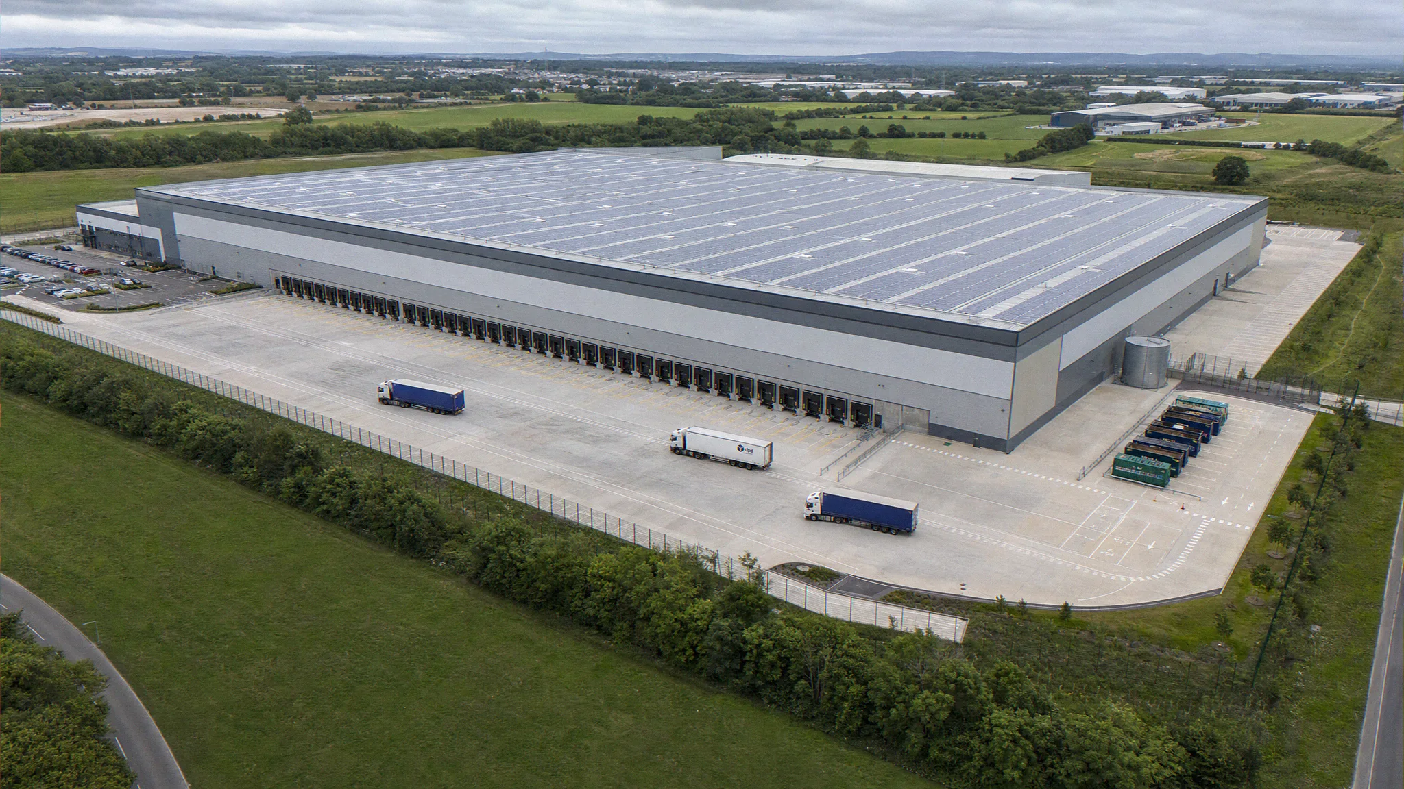 Aerial photomontage CGI of a UK big-box build-to-suit logistics park, single warehouse unit with fifty-metre yard depth, full row of dock-levellers, trailer parking, rooftop photovoltaic array, perimeter palisade, and mature landscape buffer, by StratumCGI