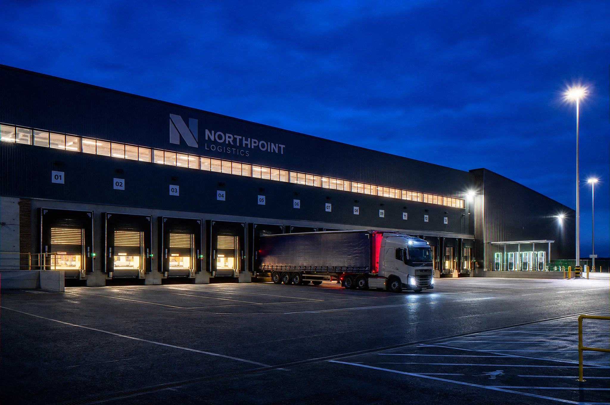Dusk photomontage CGI of a UK build-to-suit logistics warehouse dock face at blue hour, numbered dock doors with concertina shelters, articulated HGV reversing onto a leveller, EV charging stalls, and occupier wordmark on the upper facade, by StratumCGI