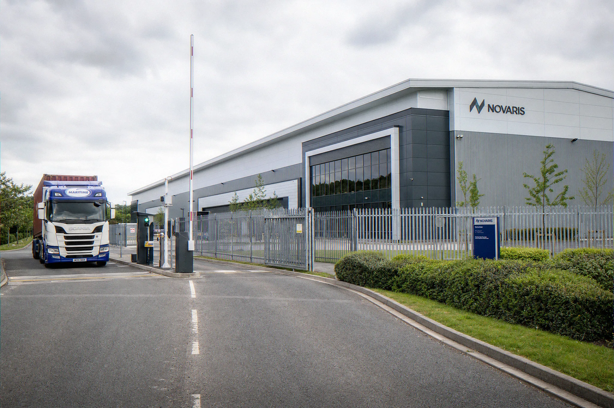 Photomontage CGI of a UK build-to-suit logistics warehouse gatehouse approach, single articulated HGV at the ANPR lane, palisade perimeter, two-storey anthracite office frontage, occupier name plate on the side elevation, by StratumCGI